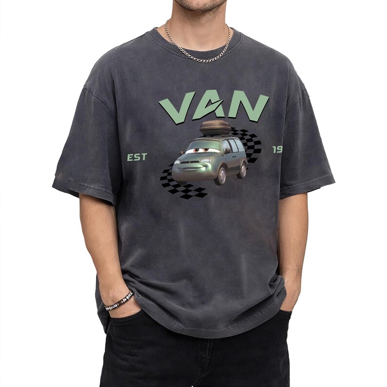 May include: Dark gray t-shirt featuring a cartoon van with the word "VAN" in light green above it. The van has a luggage carrier on top and is set against a checkered flag design. The text "EST 19" is also visible.