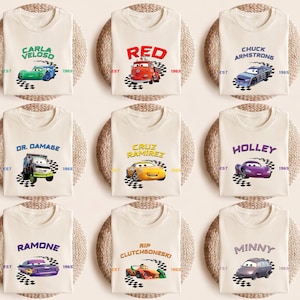 May include: Nine cream-colored t-shirts, each featuring a different character from the animated movie "Cars." The shirts display character names and car illustrations above a checkered flag design. The text "EST 1965" is also present on each shirt.