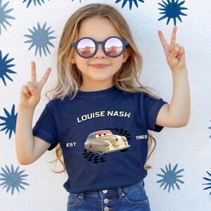 May include: A young girl wearing a navy blue t-shirt with the text "LOUISE NASH" and a cartoon car graphic. The shirt also has the text "EST 1965". She is wearing blue sunglasses and making a peace sign.