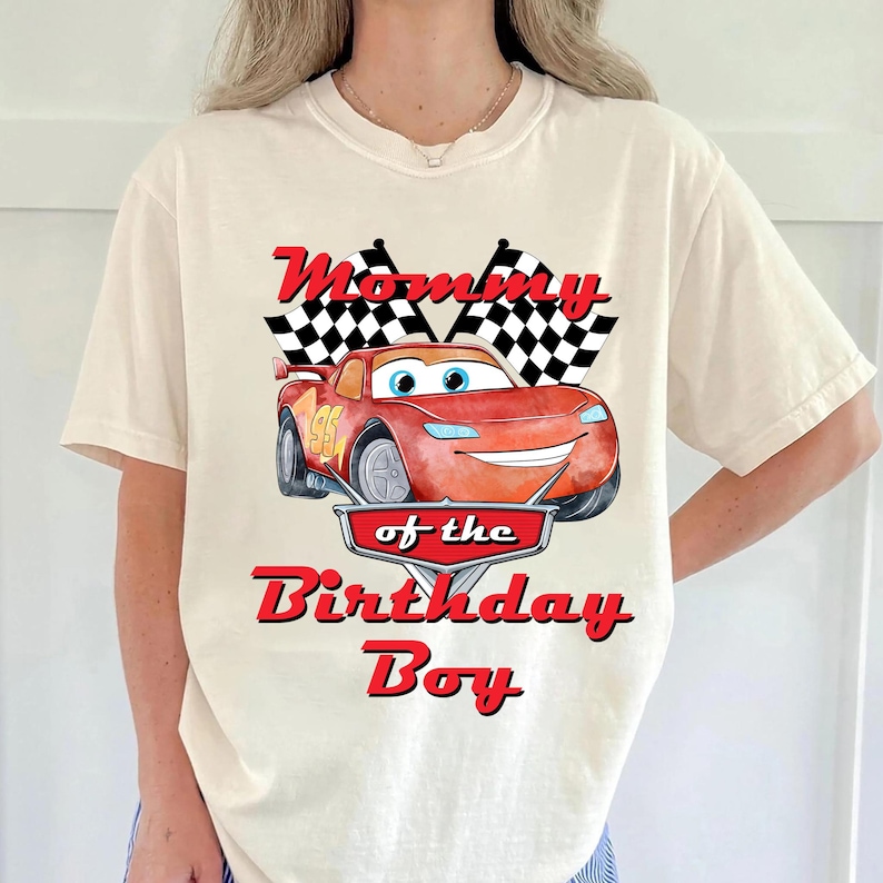 May include: Cream-colored t-shirt featuring a red cartoon race car with the number 95. The shirt has the text "Mommy of the Birthday Boy" in red. Two checkered flags are above the car.
