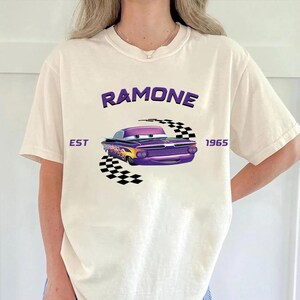 May include: Cream-colored t-shirt featuring a purple cartoon car with flames and the name "Ramone" above it. The text "EST 1965" is also printed on the shirt. A black and white checkered flag design is behind the car.