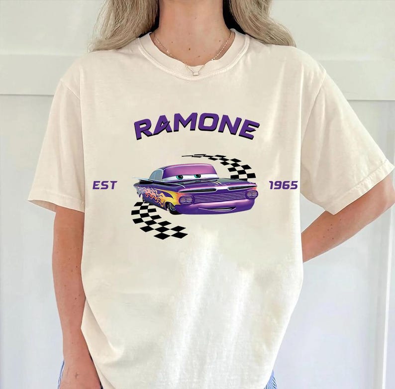 May include: Cream-colored t-shirt featuring a purple cartoon car with flames and the name "Ramone" above it. The text "EST 1965" is also printed on the shirt. A black and white checkered flag design is behind the car.