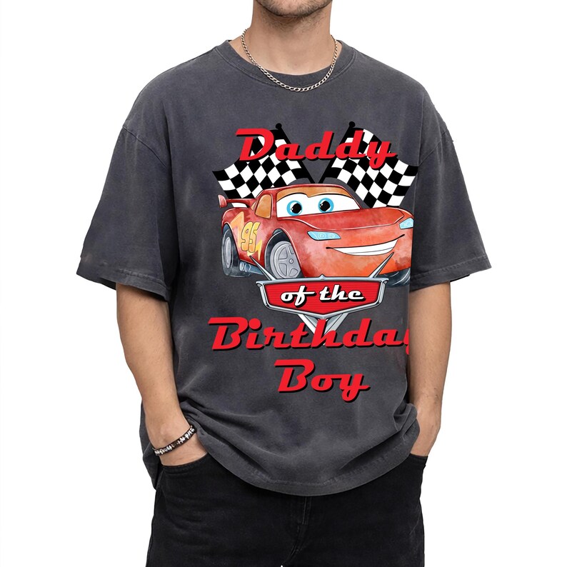 May include: Dark gray t-shirt featuring a red cartoon race car with the text "Daddy of the Birthday Boy". The design includes checkered flags and the number 95 on the car. The shirt is a relaxed fit.