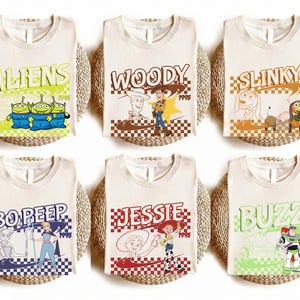 Può includere: Sei magliette color crema con personaggi di Toy Story. Ogni maglietta ha il nome di un personaggio diverso in un font retrò, con illustrazioni corrispondenti. I personaggi includono Aliens, Woody, Slinky, Bo Peep, Jessie e Buzz.