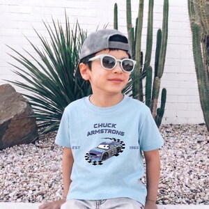 May include: A young person wearing a light blue t-shirt with the text "CHUCK ARMSTRONG" and a race car graphic. The shirt also has the text "EST 1965". They are wearing a gray cap and white sunglasses.