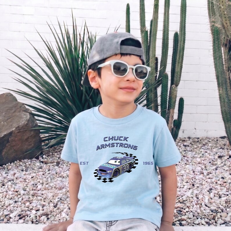 May include: A young person wearing a light blue t-shirt with the text "CHUCK ARMSTRONG" and a race car graphic. The shirt also has the text "EST 1965". They are wearing a gray cap and white sunglasses.