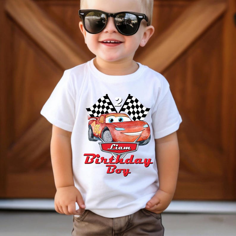 May include: White t-shirt featuring a red cartoon race car with the name "Liam" and the text "Birthday Boy" in red. Two checkered flags are above the car. The shirt is worn by a young child.