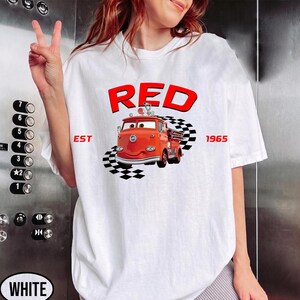 May include: White t-shirt featuring a cartoon illustration of a red fire truck with the word "RED" above it. The text "EST 1965" is also included. The fire truck is positioned on a checkered pattern.