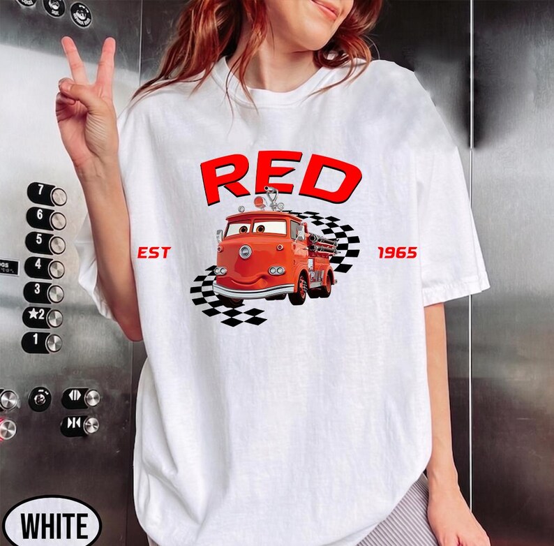 May include: White t-shirt featuring a cartoon illustration of a red fire truck with the word "RED" above it. The text "EST 1965" is also included. The fire truck is positioned on a checkered pattern.