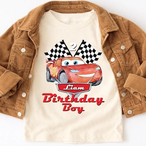 May include: A cream-colored t-shirt featuring a red cartoon race car with the number 95. The shirt has the text "Liam Birthday Boy" in red. A brown corduroy jacket is draped over the shirt.