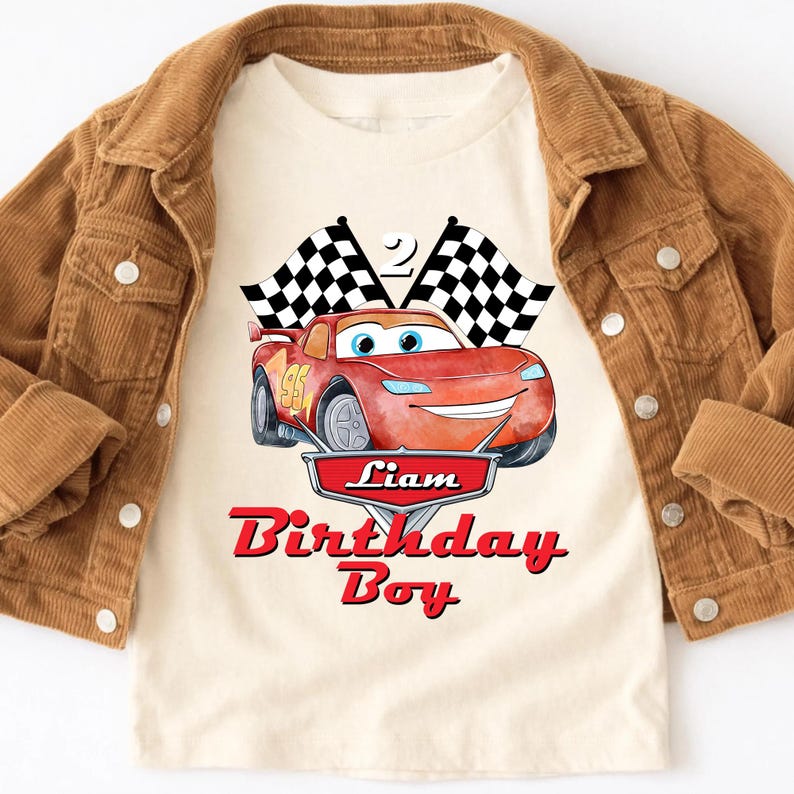 May include: A cream-colored t-shirt featuring a red cartoon race car with the number 95. The shirt has the text "Liam Birthday Boy" in red. A brown corduroy jacket is draped over the shirt.