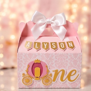 Puede incluir: Una caja de regalo rosa claro con un lazo de satén blanco en la parte superior. El frente muestra el nombre ALYSON en letras doradas, un recorte de carruaje en rosa y oro, y la palabra one en letra cursiva dorada. La caja tiene un patrón de damasco.