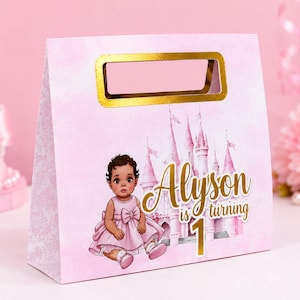 Puede incluir: Una bolsa de regalo de papel rosa claro con un asa troquelada dorada. El frente muestra una ilustración de dibujos animados de un niño pequeño con vestido rosa, un castillo rosa y el texto dorado "Alyson is turning 1". La bolsa tiene un sutil patrón de damasco.