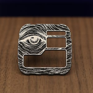 May include: A square metal pin featuring a black and silver finish with a detailed engraved eye design. The surface displays intricate swirling lines surrounding the eye, with three distinct rectangular cutouts positioned on the right side.