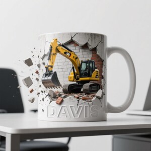 Peut inclure: Mug en c&eacute;ramique blanche avec un design de pelle m&eacute;canique jaune per&ccedil;ant un mur de briques. Le mug porte le nom "DAVIS" en relief sur le devant. La pelle porte le logo "CAT".