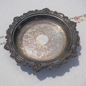 May include: A round silver-toned metal serving tray featuring an intricate embossed floral and scrollwork pattern across the base. The tray has a wide, scalloped rim with decorative shell-like motifs and a polished, reflective center surface.