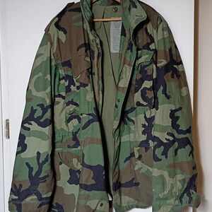 May include: A field jacket featuring a woodland camouflage pattern in shades of dark green, light green, brown, and black. The jacket includes a high collar, a front zipper closure, two chest pockets with flaps, and two lower pockets with flaps.