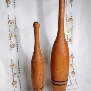 May include: Two light brown wooden exercise clubs of different heights. Each club features a rounded top, a long slender neck with horizontal grooved detailing, a wider bulbous body, and two thin black horizontal rings near the flat base.