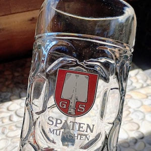 May include: A clear glass beer mug featuring a dimpled texture and a printed logo. The logo displays a red shield with a white shovel icon, the letters G and S, and the text SPATEN MÜNCHEN printed in black lettering below the shield design.