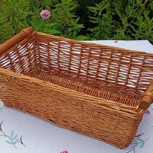 May include: A rectangular woven wicker basket featuring a light brown natural finish. The basket has an open-top design with a sturdy, smooth wooden handle attached to each of the two shorter ends, secured by thin, flexible woven wicker strips.