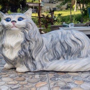 May include: A ceramic cat figurine featuring long, flowing fur painted in shades of grey, white, and charcoal. The cat has bright blue eyes, a pink nose, and is depicted in a resting, lying-down position with its head turned slightly to the side.