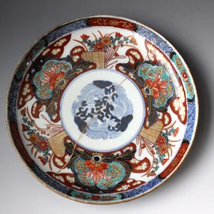 May include: A round ceramic plate featuring a central light blue floral motif surrounded by intricate red, orange, green, and dark blue patterns. The plate displays a visible thin gold-colored crack running across the surface from top to bottom.