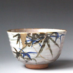以下が含まれることがあります： 白地に青と濃い緑色の竹の葉が手描きされた陶器のボウル。ボウルの側面には、金継ぎの修復技法に特徴的な、垂直に走る目立つ金色の修復線が施されています。