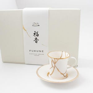 以下が含まれることがあります： 金継ぎ風の金色のひび割れ模様が施された白い陶器のカップとソーサーのセット。セットは白い長方形の箱に入っており、紙の帯には「福音」という文字と「FUKUNE KINTSUGI BRAND JAPAN」というテキストが印刷されています。