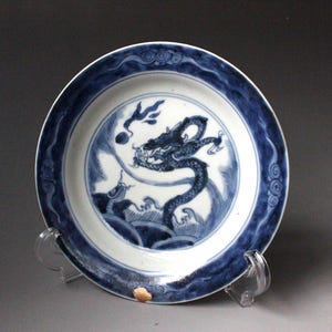 Puede incluir: Un plato redondo de cerámica con un motivo de dragón pintado a mano en azul y blanco en el centro. El borde está decorado con un patrón azul ondulado. El plato se exhibe sobre un soporte de plástico transparente, mostrando el diseño detallado.