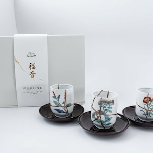 May include: Three white ceramic tea cups with floral patterns and gold-colored kintsugi-style cracks, each resting on a dark brown saucer. A white gift box with the text "FUKUNE" and Japanese characters is positioned behind the cups.
