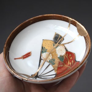 May include: A white ceramic bowl featuring a gold-painted rim and a central fan-shaped graphic in red, black, and gold. The bowl displays visible gold-filled cracks across the surface, highlighting a repair technique that accentuates the fractures.