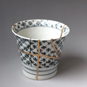 May include: A white ceramic cup featuring a blue and black grid pattern with small floral motifs. The cup displays visible cracks repaired with gold lacquer, highlighting the kintsugi technique across the exterior and the rim.