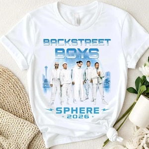 Könnte beinhalten: Ein weißes kurzärmeliges T-Shirt mit einem Grafikdruck der Backstreet Boys in weißen Outfits vor einem blauen Stadtbild-Hintergrund. Der Text lautet BACKSTREET BOYS oben und SPHERE 2026 unten in einer blauen Verlaufsschrift.