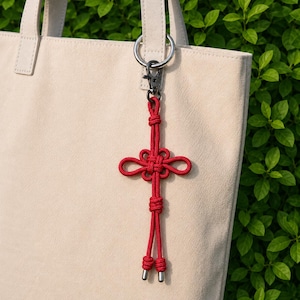Könnte beinhalten: Ein roter chinesischer Knoten-Schlüsselanhänger mit dekorativem Design, befestigt an einem silbernen Schlüsselring und einer beigen Tragetasche. Der Knoten besteht aus gewebtem Kordel und hat ein symmetrisches Muster.