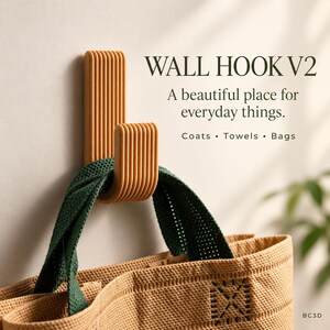 May include: An amber-colored wall hook with a ribbed, vertical design. The text reads: WALL HOOK V2, A beautiful place for everyday things. Coats • Towels • Bags. A tan bag with dark green handles hangs from the hook, which is mounted on a wall.