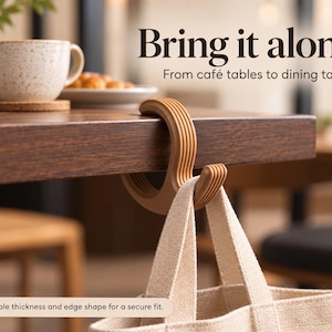 May include: A light brown, C-shaped bag hook with a ribbed texture, hanging from the edge of a wooden table. The text "Bring it along. From café tables to dining tables" and "Check table thickness and edge shape for a secure fit" is visible.