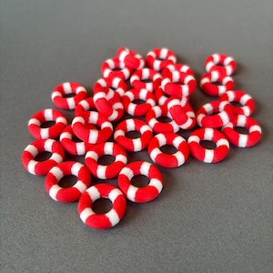 May include: A collection of small, red and white striped life rings. These miniature life rings are arranged on a grey surface. The life rings are a nautical theme.