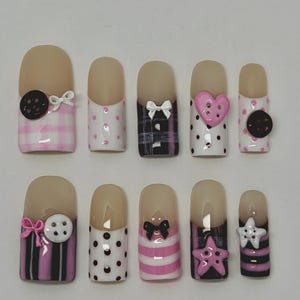 May include: A set of ten artificial nails with various designs. The nails feature a nude base with decorative elements such as bows, polka dots, hearts, and cookies. Colors include pink, black, white, and brown. Some nails have a plaid or striped pattern.