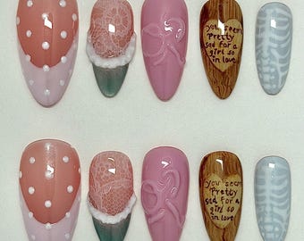 May include: A set of ten press-on nails featuring diverse designs: pink with white polka dots, lace patterns, raised ribbon motifs, wood grain textures with the text 'you seem pretty sad for a girl so in love', and light blue skeleton rib cage art.