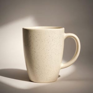 May include: Cream-colored ceramic mug with a speckled pattern. The mug has a curved handle and a cylindrical shape. The mug is approximately 4 inches tall and 3 inches in diameter.