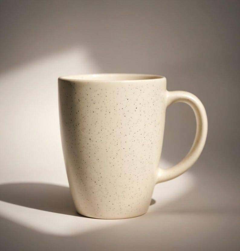 May include: Cream-colored ceramic mug with a speckled pattern. The mug has a curved handle and a cylindrical shape. The mug is approximately 4 inches tall and 3 inches in diameter.