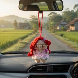 May include: A handmade crochet doll with red hair and a pink dress hangs from a car's rearview mirror. The doll is suspended by a red string. The car's dashboard and windshield are visible, with a scenic road and fields in the background.