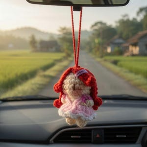 May include: A handmade crochet doll car charm. The doll has red hair, a pink dress, and a white skirt. It hangs from a red string, suspended from a car's rearview mirror. The background shows a road and green fields.