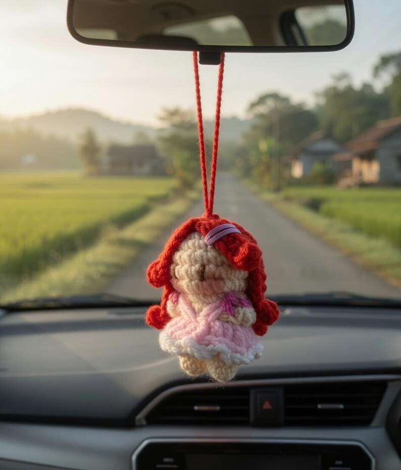 May include: A handmade crochet doll car charm. The doll has red hair, a pink dress, and a white skirt. It hangs from a red string, suspended from a car's rearview mirror. The background shows a road and green fields.