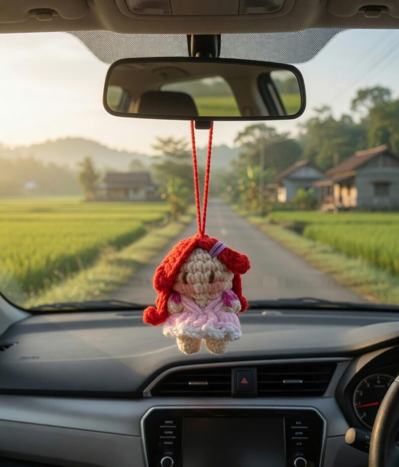 May include: A handmade crochet doll with red hair and a pink dress hangs from a car's rearview mirror. The doll is suspended by a red string. The car's dashboard and windshield are visible, with a scenic road and fields in the background.