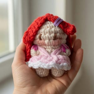 May include: A small, handmade crochet doll with red hair, a pink dress, and a white trim. The doll has a light skin tone and is held in a hand. The doll's hair is styled with a purple barrette. The doll's dress has pink accents.