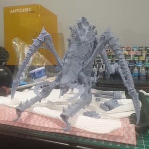 Puede incluir: Un modelo gris detallado de una criatura grande con forma de insecto con múltiples patas y apéndices afilados. El modelo está sobre una mesa con otros objetos, incluyendo una caja amarilla con la palabra "ANYCUBIC". La criatura parece ser un modelo impreso en 3D.