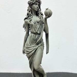Puede incluir: Una escultura gris de una mujer con una corona floral y un vestido drapeado, sosteniendo una fruta. La estatua tiene cabello largo y suelto y está de pie sobre una base texturizada. La pose y el atuendo de la figura sugieren un tema clásico o mitológico.