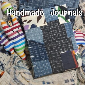 Può includere: Un diario rettangolare con una copertina patchwork realizzata con vari tessuti in denim blu, motivi a griglia e stampe floreali, con il testo "Handmade Journals" in alto, accompagnato da piccole figure di animali in tessuto imbottito a righe arcobaleno.