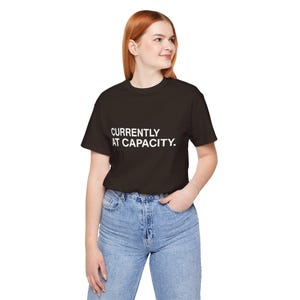 May include: A black t-shirt with the white text "CURRENTLY AT CAPACITY." The shirt is short-sleeved and worn with blue jeans. The person is wearing a silver bracelet.